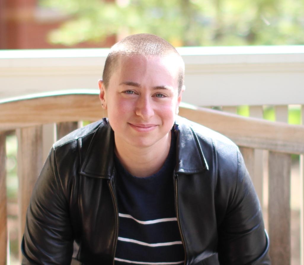 A White non-binary person with a buzzcut smiles at the camera.
